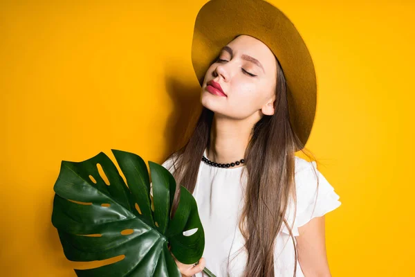 Menina modelo atraente confiante com cabelos longos mantém uma folha verde, na cabeça um grande chapéu de moda posando em um fundo amarelo — Fotografia de Stock