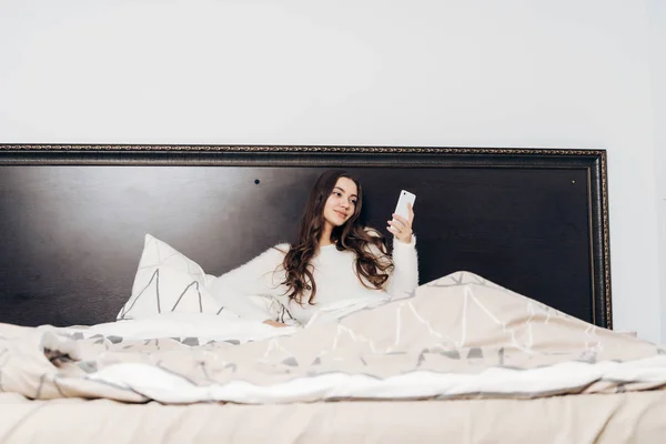 Menina sonolenta bonito em pijama branco encontra-se na cama tarde da noite, descansando depois de um dia de trabalho duro, olha para um smartphone — Fotografia de Stock