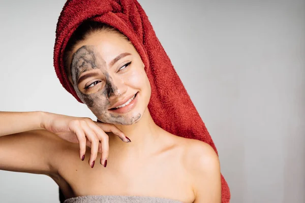 Belle jeune fille avec une serviette rouge sur la tête a appliqué un masque d'argile sur la moitié de son visage, souriant — Photo