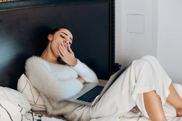 Уставшая сонная девушка фрилансер в пижаме сидит в постели, работает на ноутбуке поздно ночью — стоковое фото