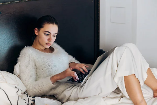 Серйозна молода дівчина в білій піжамі сидить в ліжку пізно вночі, відпочиває і дивиться серіали на її ноутбук — стокове фото