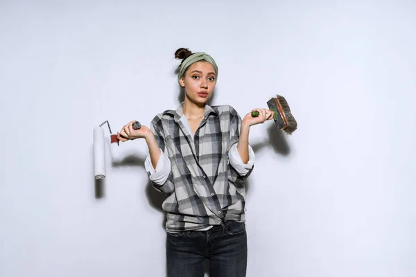 Удивлена молодая девушка в клетчатой рубашке делает ремонт в своей квартире, держа кисть и ролики для покраски стен и не может выбрать — стоковое фото