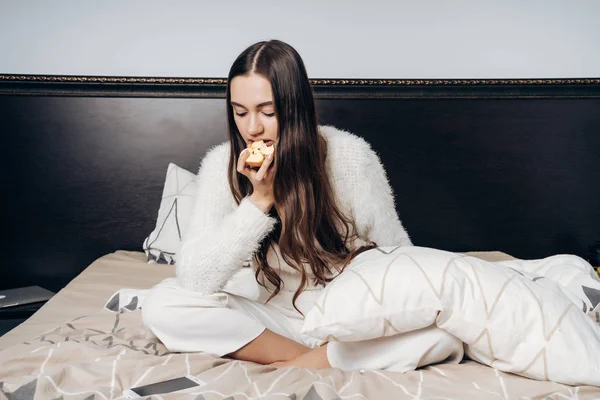 Sonolenta menina de cabelos longos em pijama branco acordou de manhã cedo, senta-se na cama e come uma maçã no café da manhã — Fotografia de Stock