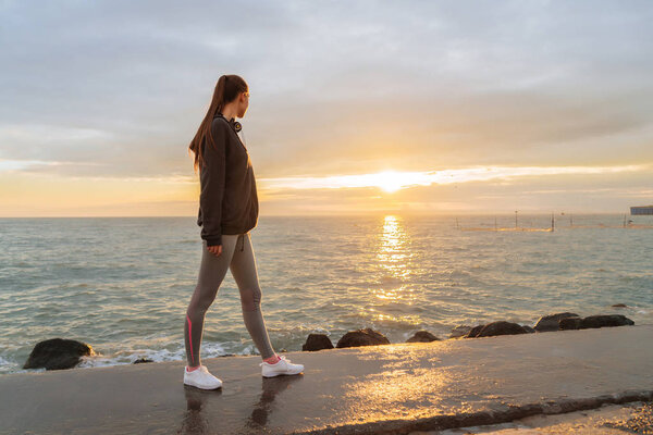 a slender tall girl in sports clothes rests after jogging at the sea at sunset, listens to the music in the headphones