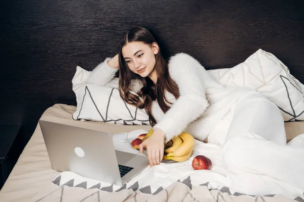 Hermosa joven se acuesta en una cama por la noche, descansando después de un día de trabajo y viendo una película interesante en su computadora portátil — Foto de Stock