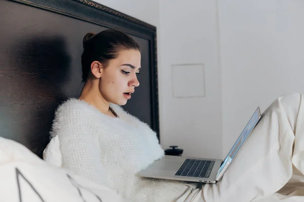 Triste joven se acuesta en la cama tarde en la noche, bajo una manta blanca, viendo series de televisión en su computadora portátil —  Fotos de Stock