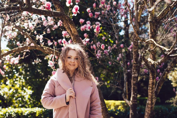 Красива кучерява жінка в рожевому пальто позує в парку, дивлячись на камеру і посміхаючись — стокове фото