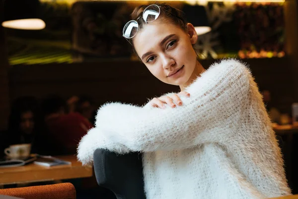 Очаровательная студентка в белой стильной блузке сидит в ресторане, смотрит в камеру и улыбается — стоковое фото