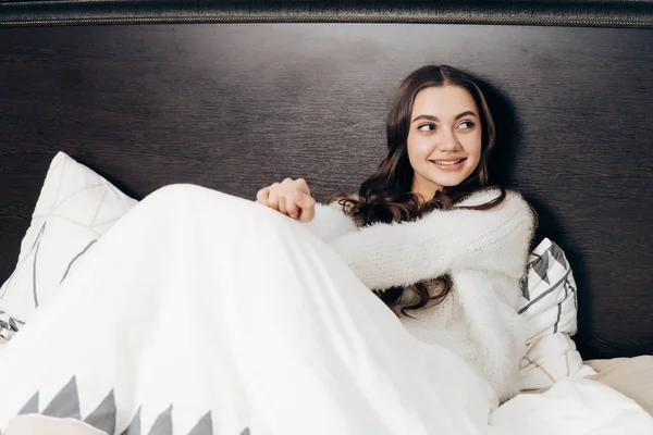 Encantadora jovem encontra-se na cama sob um cobertor branco, bom humor, sorrindo — Fotografia de Stock