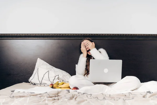 Carino ragazza in pigiama bianco si siede a letto, guarda film divertente sul suo computer portatile, coperto il viso con le mani — Foto Stock