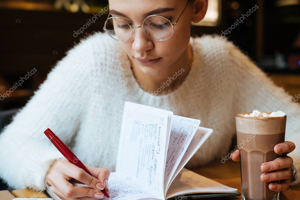 serious young girl freelancer with glasses works, writes in a notebook and drinks a hot latte in a cafe