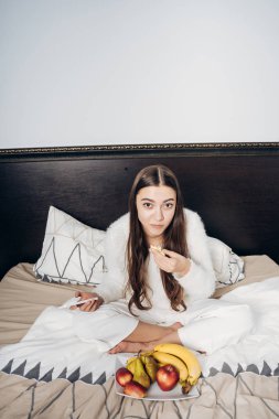 uzun saçlı kız beyaz pijamalı yatakta meyve yeme erken oturur.