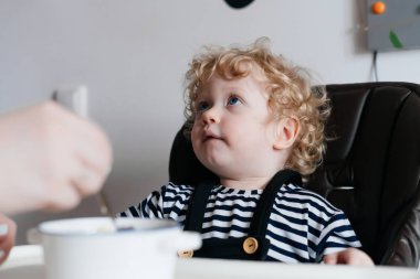Sevimli sarışın kıvırcık yürümeye başlayan çocuk mutfakta oturuyor, Anne onu yararlı bir lapa beslemeleri