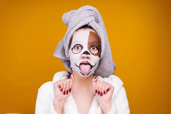 A funny young girl with a towel on her head and a bathrobe showing her tongue, and a mask with a muzzle of a dog — Stock Photo, Image