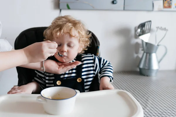 Заботливая мать кормит своего маленького кудрявого сына полезным вкусным супом, семейным ужином — стоковое фото