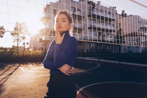 Confiada chica rubia con estilo en un suéter azul de moda posando en un campo de deportes en los rayos del sol de la mañana — Foto de Stock