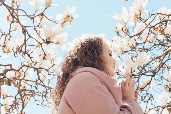 zarif kıvırcık kadın parkta mavi gökyüzünün altında çiçek açan bir Manolya koklama