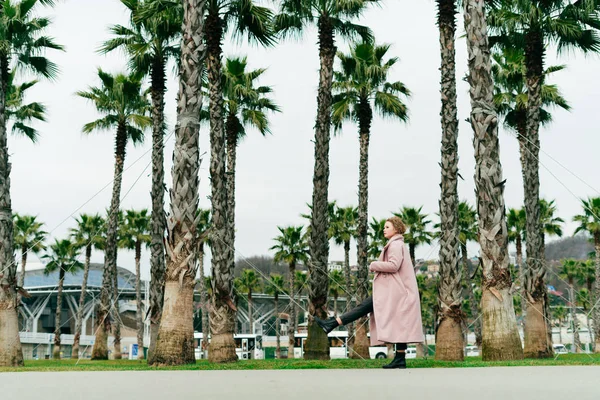 Stilig ung dame i lang rosa frakk som går ved siden av høye, grønne håndflater – stockfoto