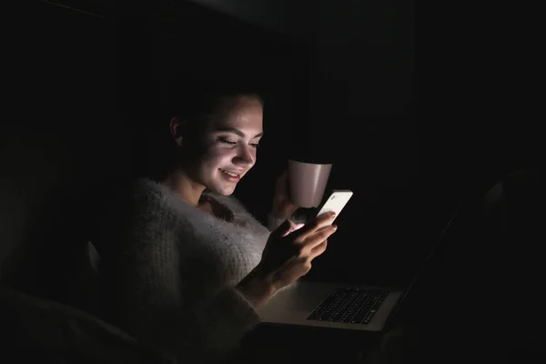 Bella ragazza sorridente seduta a letto a tarda notte, bevendo tè e scrivendo un messaggio sul suo smartphone — Foto Stock