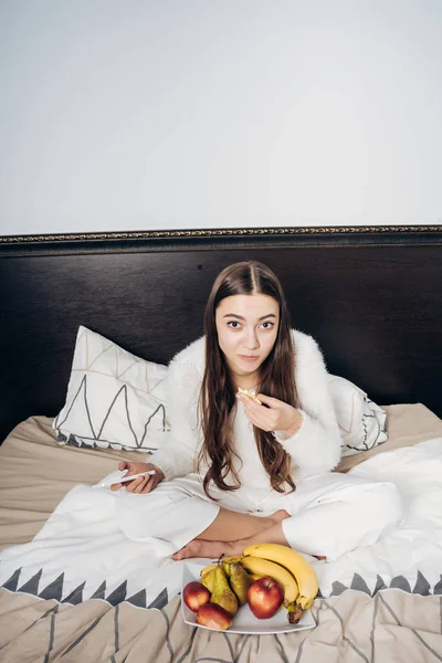uzun saçlı kız beyaz pijamalı yatakta meyve yeme erken oturur.