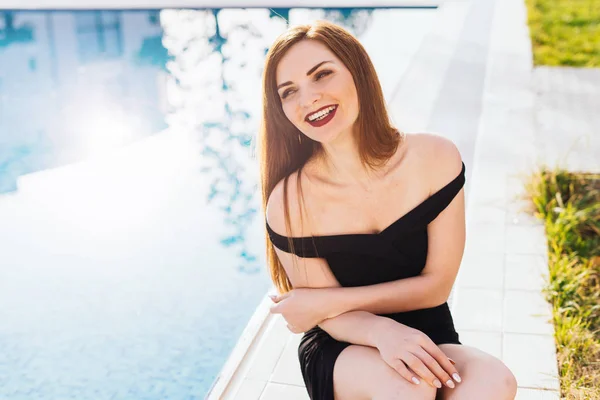 Uma luxuosa mulher de cabelos longos em um vestido preto descansa à beira da piscina azul em seu jardim ao sol, ri — Fotografia de Stock