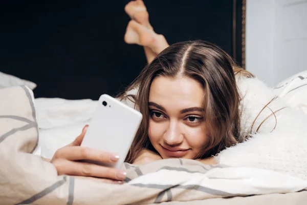 sleepy sweet girl in white pajamas wakes up early, lies in bed and writes a message on her smartphone