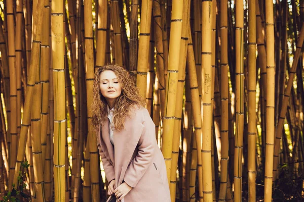 Curly young woman in a stylish pink coat posing against a bamboo tree in a park — Stock Photo, Image