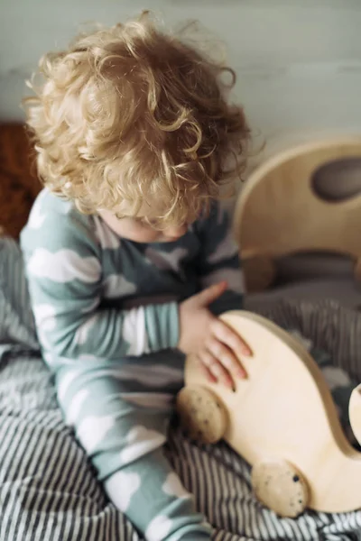 Маленька кучерява блондинка малюк у блакитній піжамі сидить на ліжку, граючи з дерев'яною іграшкою — стокове фото