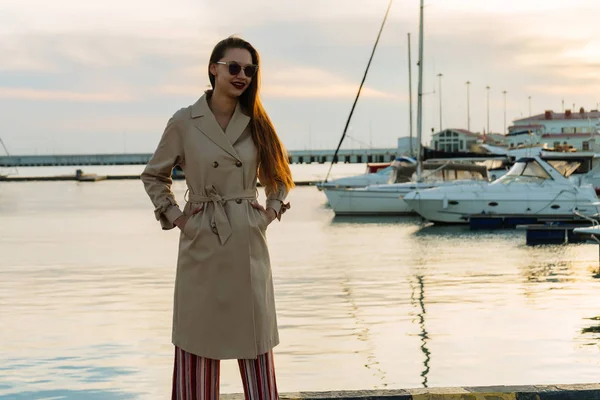 Lujosa chica de pelo largo en gafas de sol y abrigo de moda posando en el puerto, sonriendo — Foto de Stock