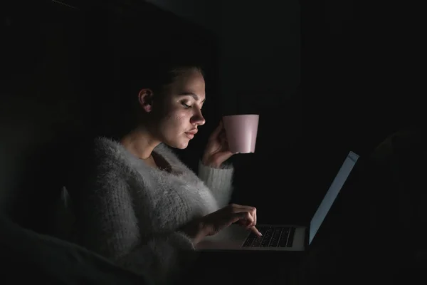 Серйозна молода дівчина фрілансер сидить у ліжку пізно вночі, п'є чай і працює на ноутбуці, в темряві — стокове фото