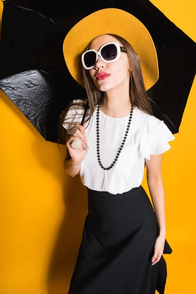 Luxuosa menina bonita em óculos de sol e chapéu elegante posando no fundo amarelo, segurando guarda-chuva, estilo — Fotografia de Stock