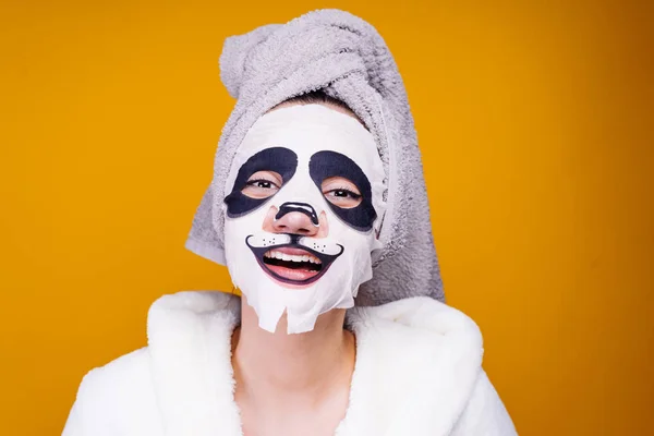 Весела молода дівчина з рушником на голові посміхається, піклується про себе, на її обличчі зволожуюча маска з обличчям панди — стокове фото