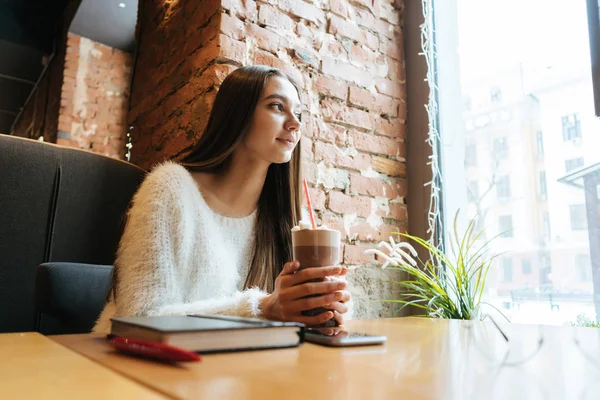 Hermosa chica de pelo largo en un suéter blanco descansando en un café, bebiendo un delicioso café con leche y mirando por la ventana —  Fotos de Stock