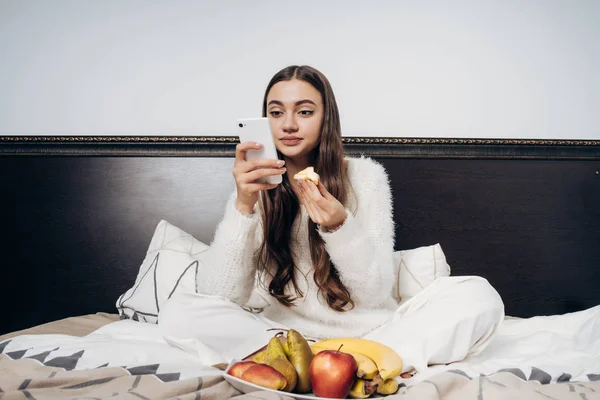 Bonito de cabelos longos menina senta-se na cama tarde da noite, come frutas e olha para o seu smartphone — Fotografia de Stock