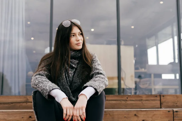 Beautiful dark-haired girl in a trendy gray coat sits on the steps and rests, smiling — Stock Photo, Image