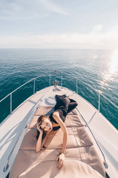 An attractive young woman in black summer clothes lies on her yacht, enjoys vacation and sea — Stock Photo, Image