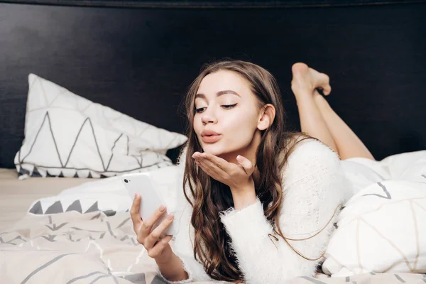 Vacker ung flicka ligger i sängen, håller smartphone och skickar en air kyss — Stockfoto