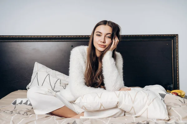 Encantadora jovem em pijama branco sentado na cama de manhã cedo, sorrindo e olhando para a câmera — Fotografia de Stock