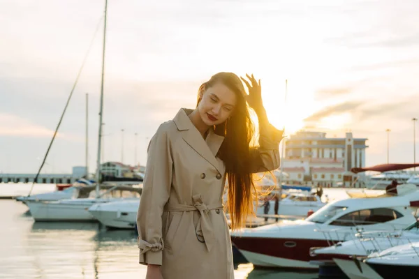Atrakcyjny dziewczynka długowłosy w stylowy płaszcz beżowy czeka na jej jacht w morskim, uśmiechając się — Zdjęcie stockowe