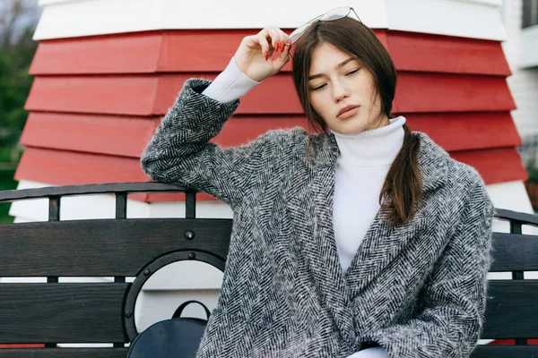 Élégante fille aux cheveux bruns dans un manteau gris à la mode assis sur un banc à l'extérieur — Photo