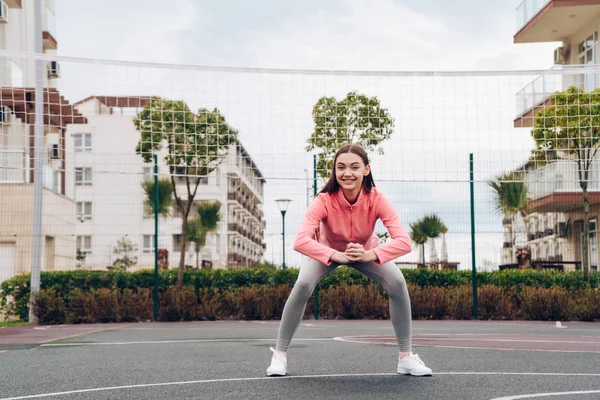 Actieve lachende meisje in een roze trui treinen en kniebuigingen op een sportveld — Stockfoto