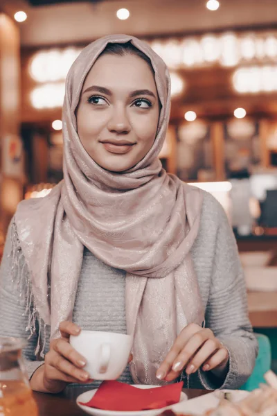 Привлекательная молодая девушка с платком на голове обедает в уютном ресторане, думает о чем-то — стоковое фото