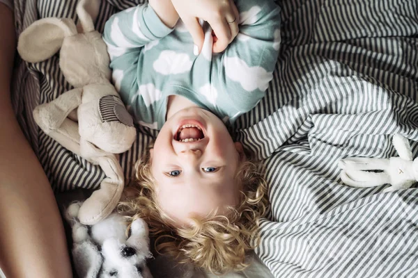 Блондин, кудрявый мальчик в голубой пижаме лежит на кровати рядом со своей матерью, смеется — стоковое фото