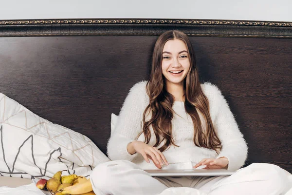 Riendo chica de pelo largo en una chaqueta blanca se despertó temprano en la mañana, se sienta en la cama y come fruta — Foto de Stock