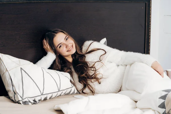 Γοητευτικό κορίτσι με μακριά μαλλιά είναι ανάπαυση στο κρεβάτι το βράδυ, σύμφωνα με μια άσπρη κουβέρτα — Φωτογραφία Αρχείου