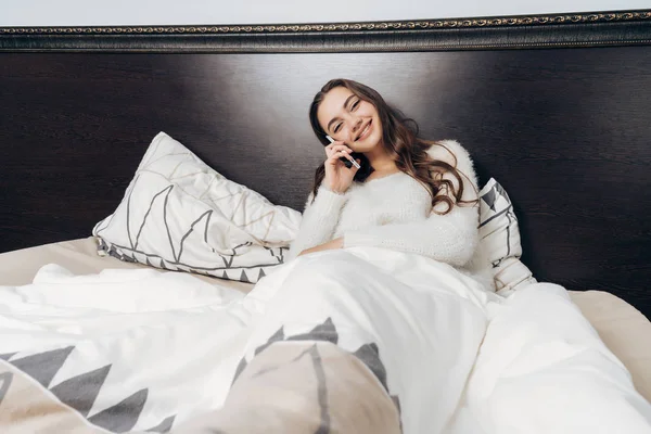 Encantadora chica encantadora en pijama blanco se encuentra en la cama temprano en la mañana, hablando por teléfono y sonriendo — Foto de Stock