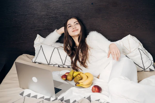 Chica de pelo largo soñadora en pijama blanco descansando en la cama, viendo la película en su computadora portátil y comiendo fruta — Foto de Stock