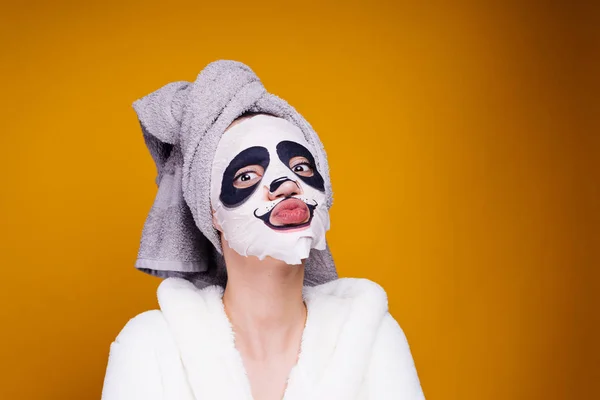 Ένα αστείο κορίτσι με μια πετσέτα στο κεφάλι της και μια άσπρη ρόμπα φροντίζει τον εαυτό της, στο πρόσωπό της μια μάσκα με πρόσωπο panda — Φωτογραφία Αρχείου