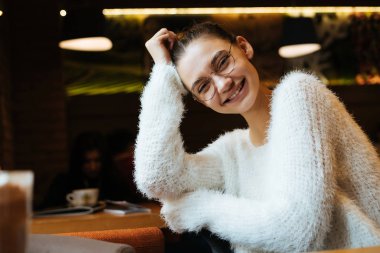 Sevimli Genç kız öğrenci bir beyaz kazaklı ve gözlük oturur bir restoran ve onu yemek için bekler güler