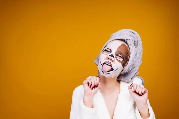 Una joven divertida con una túnica blanca y con una toalla en la cabeza muestra la lengua, en su cara una máscara hidratante con la cara de un perro — Foto de Stock